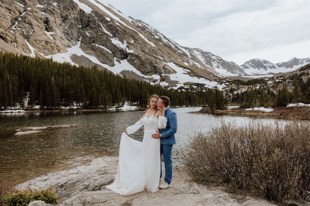 bride and groom embracing in front of an alpine lake with mountains in the background