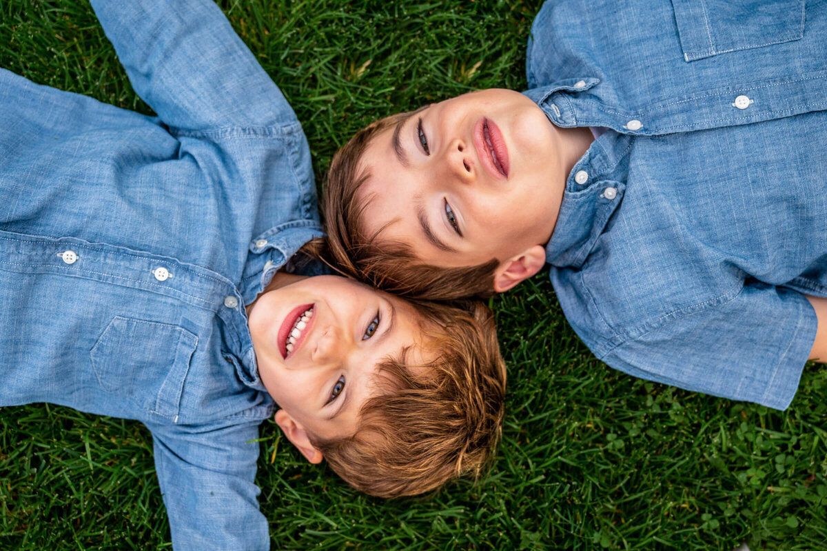 Two young boys laying on grass, heads together, opposite each other.