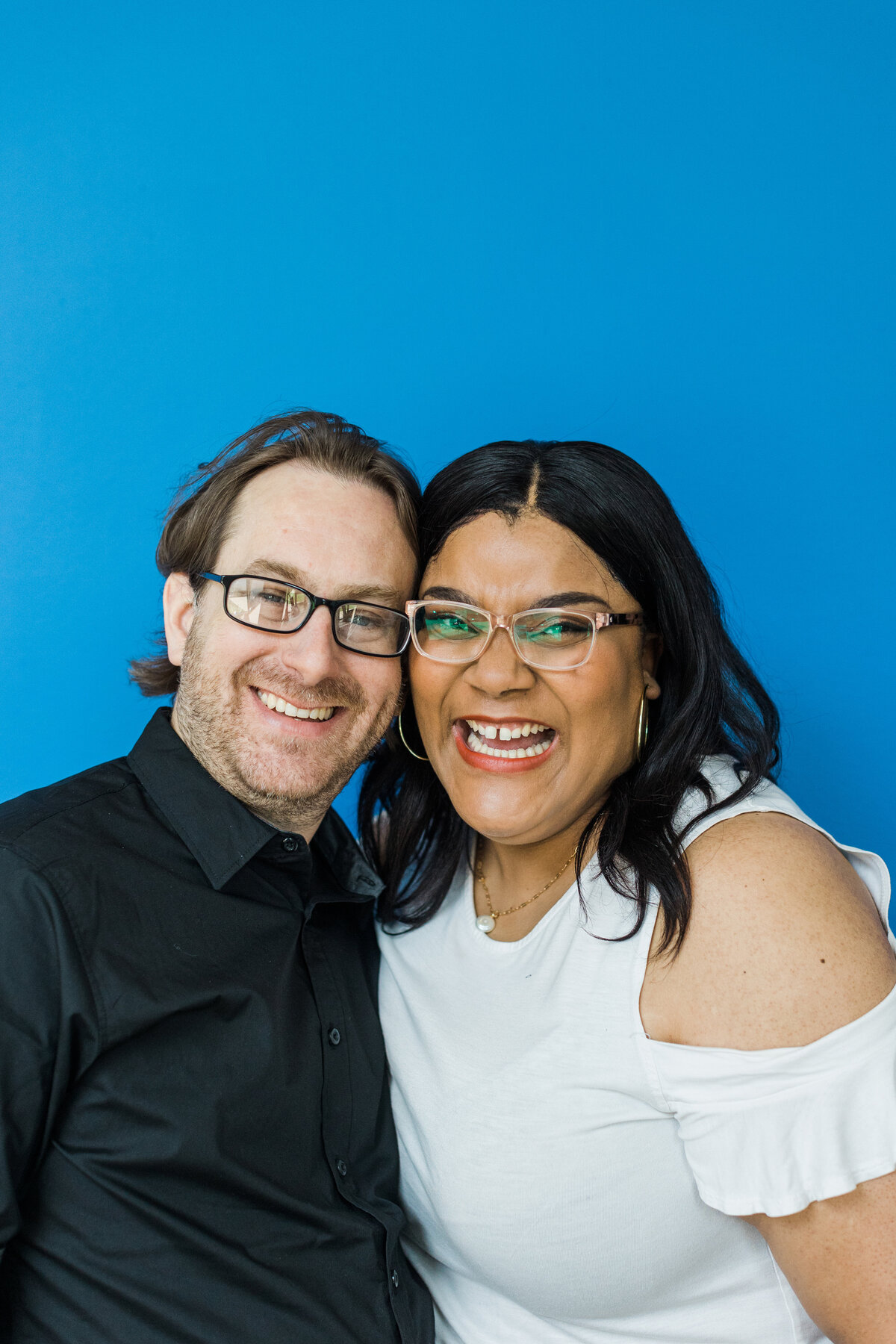 A couple joyfully smiling and posing with their faces touching in front of a blue background during their studio engagement session in Dallas, Texas. The woman on the right is wearing a white top and glasses while the man on the left is wearing a black dress shirt and glasses.