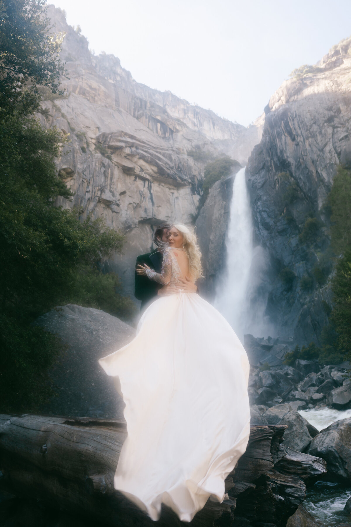Closeup of bride and groom eloping at Yosemite waterfall at golden hour with her wind and Claire Pettibone gown flowing dramatically in the wind and a slight haze in the lens from the water