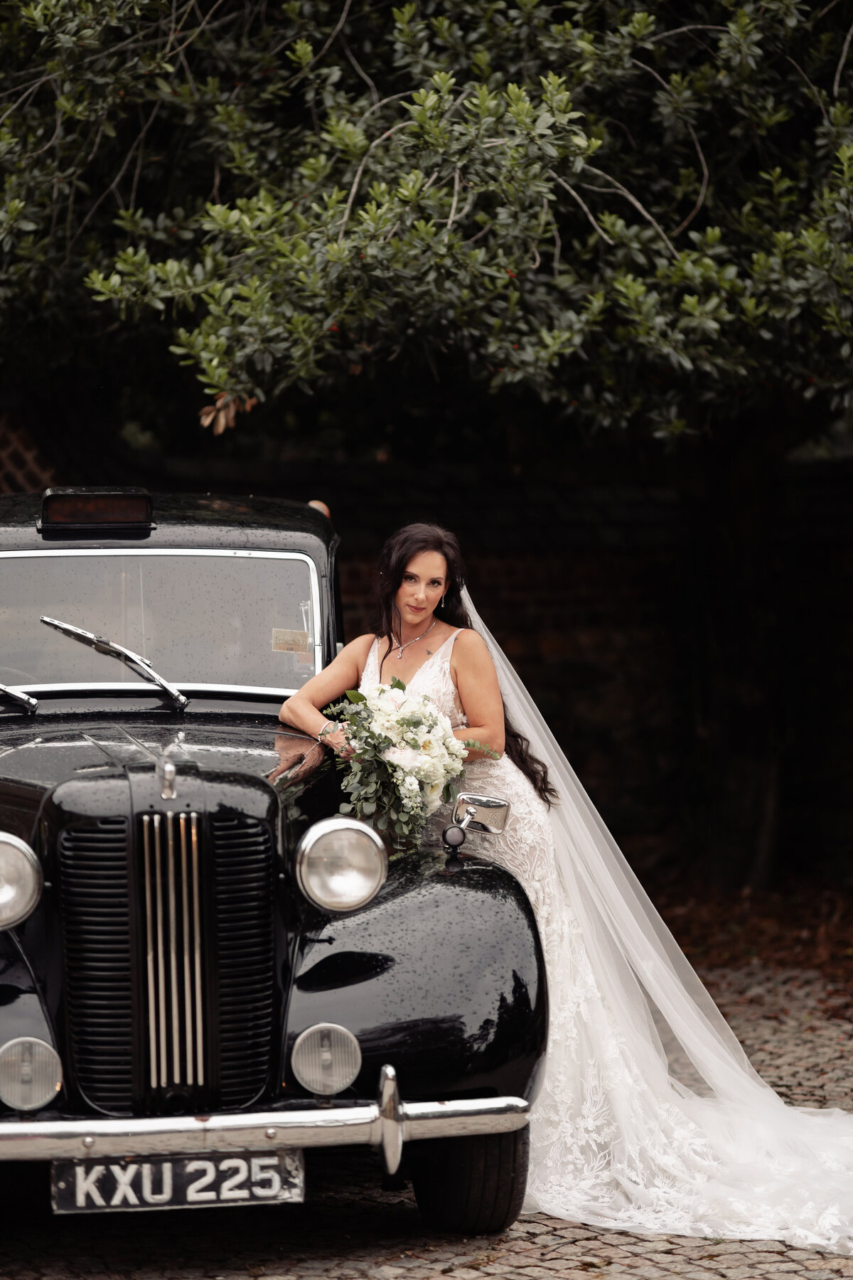 Bride leaned up against an old car with her bouquet draped across it