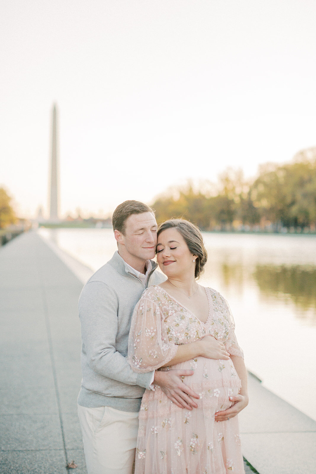 Pregnant mother in pink Needle & Thread dress leans into her husband as they stand by the Reflecting Pool in DC.