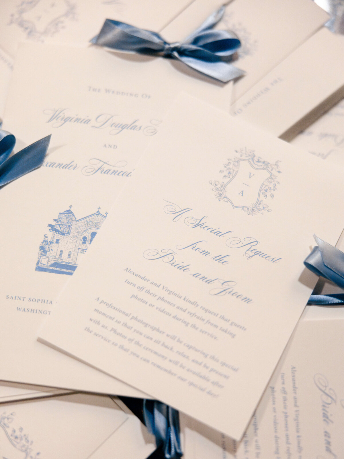 A stack of elegant wedding invitation cards features blue bows and ornate printed details. The invitations request guests to refrain from using mobile devices during the ceremony and ask for professional photographer preference.