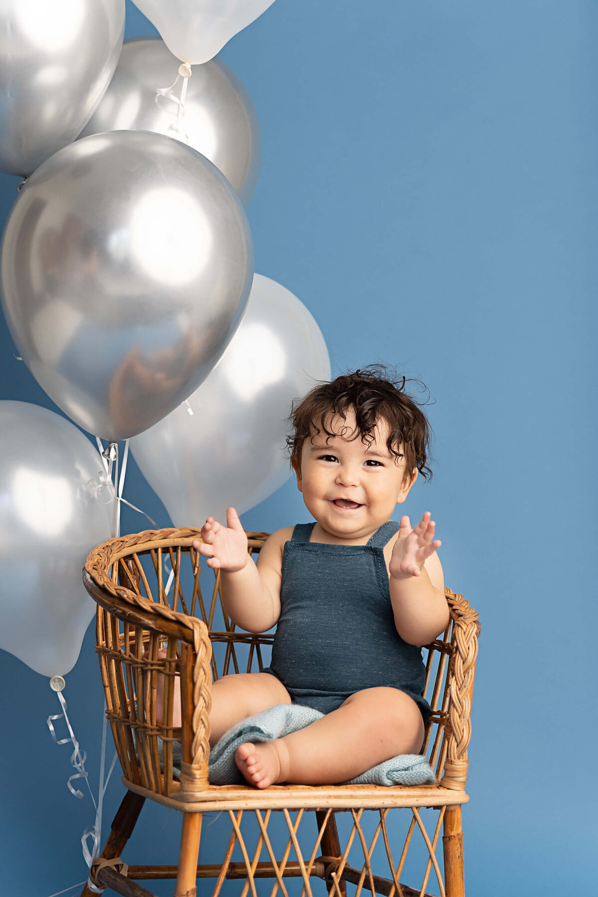 baby boy sitting in little chair smiling and clapping with silver balloons in the backgound and blue backdrop