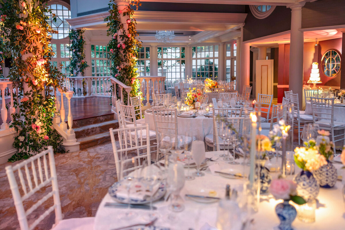 An elegantly decorated event hall with white tables and chairs, adorned with floral centerpieces and soft lighting. The room features large windows, plants climbing columns, and a staircase, creating a romantic and inviting atmosphere.