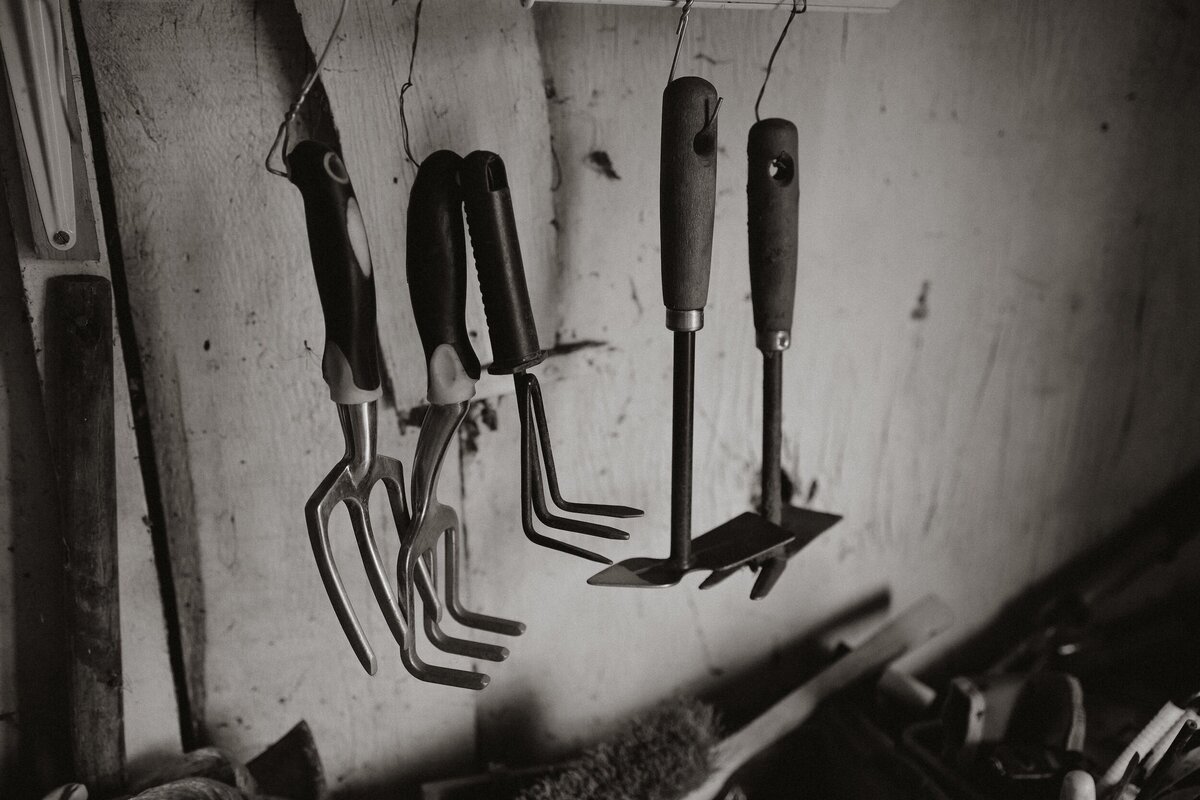 Black and white photograph of gardening tools hanging in a shed, part of The Quiet Gift series by Marie Kenny.