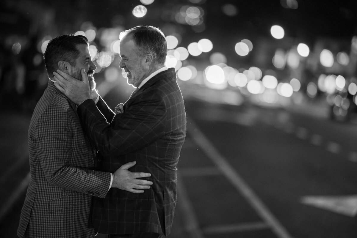 Two grooms in suits embrace and smile at each other on a city street at night, with blurred lights in the background. Black and white image. This photo was taken by a NYC wedding photographer for rooftop ceremonies