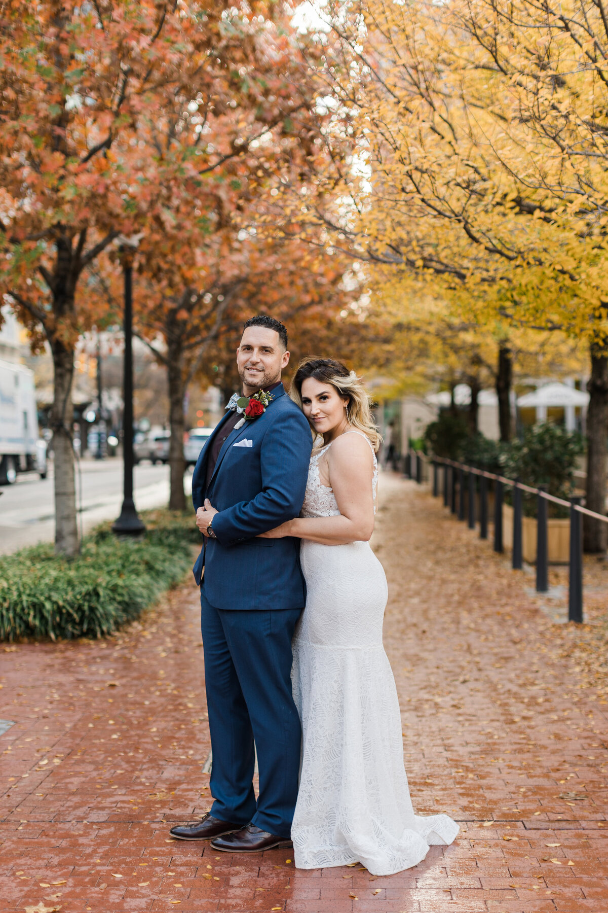 Portrait of a bride and groom posing in downtown Fort Worth after their elopement ceremony at the Tarrant County Courthouse in Fort Worth, Texas. The bride is "the big spoon" while the groom is " the little spoon," and they are backed by many fall colors on the trees that frame the scene. The bride is on the right and is wearing a sleeveless, white dress. The groom is on the left and is wearing a navy suit with a boutonniere.
