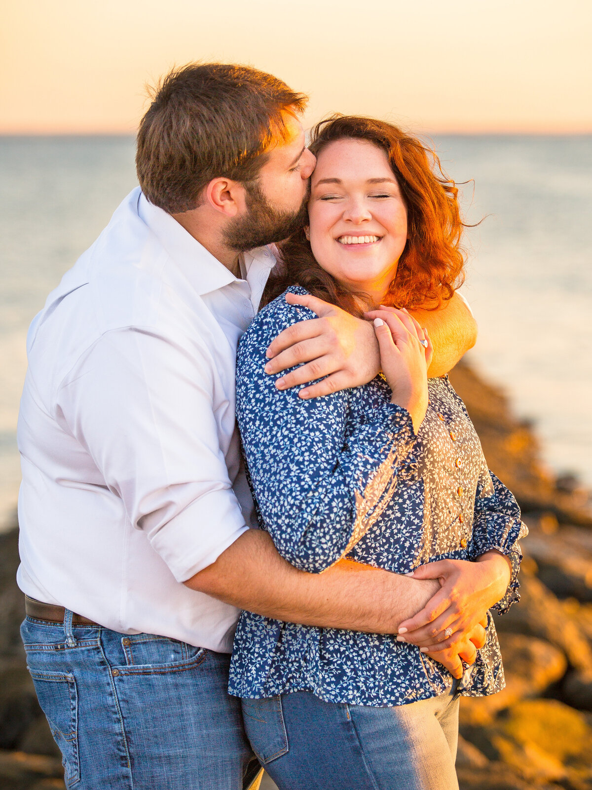 Woman laughs as her fiancé hugs her from behind and kisses her cheek during their engagement session.
