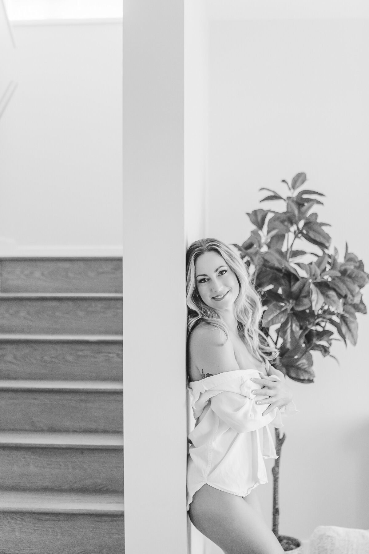 A woman leaning against the wall near a staircase, smiling warmly while wearing an oversized shirt in a bright, airy space.