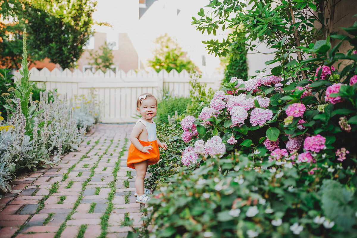 Baby girl in orange shorts laughing in a garden with pink flowers in Fells Point Baltimore Maryland