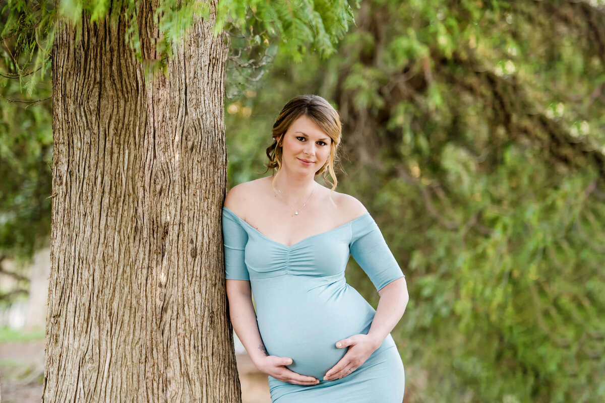 pregnant woman cradling her belly leaning against a tree in a blue maternity dress for her outdoor maternity photos in Burlington, Ontario