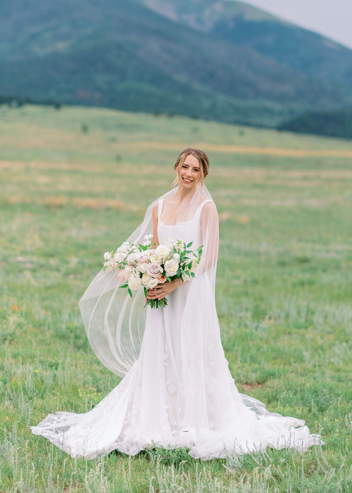 beautiful bride in a flowing white dress smiles at the camera while the wind blows her delicate veil around her shoulders