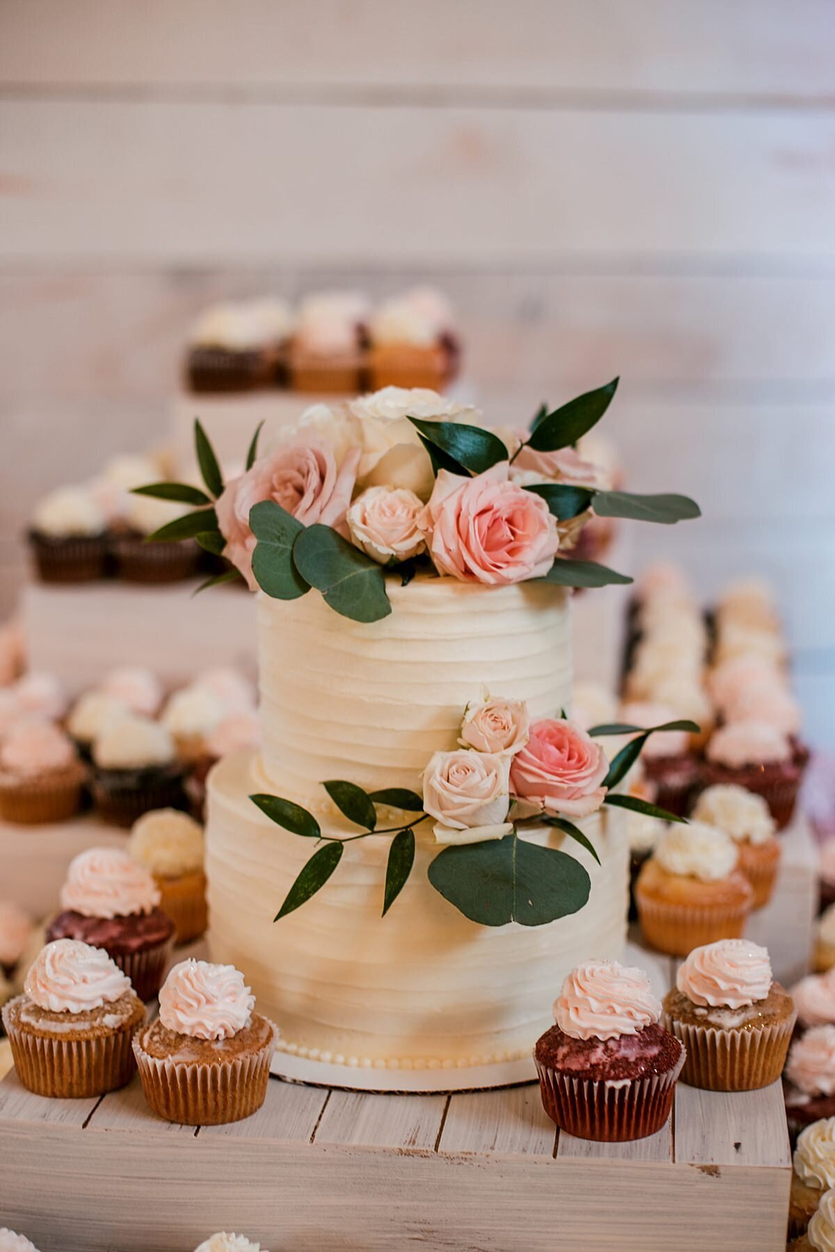 A small two tier wedding cake decorated with pink roses, blush roses and greenery sits among assorted cupcakes with rosettes of buttercream on top.