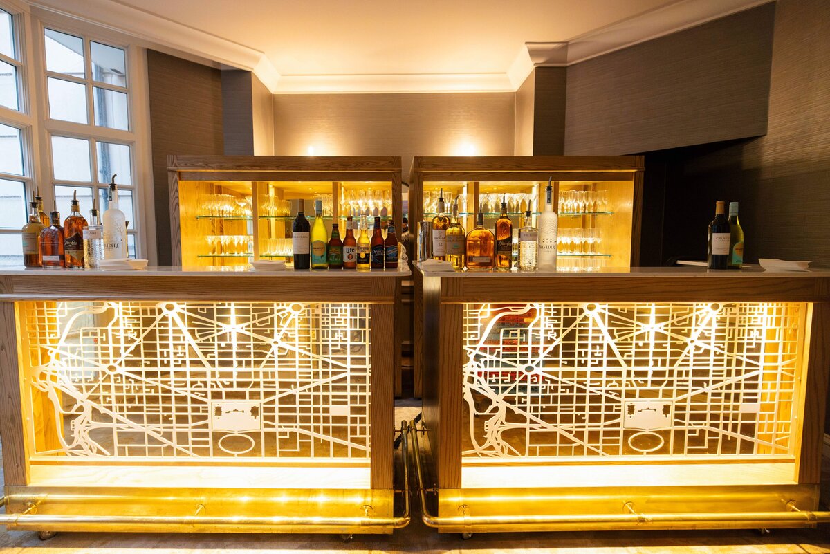 A stylish bar with two illuminated counters displaying various bottles of liquor. Behind the counters, shelves with more bottles are visible. The room has a warm, cozy ambiance with soft lighting and large windows.