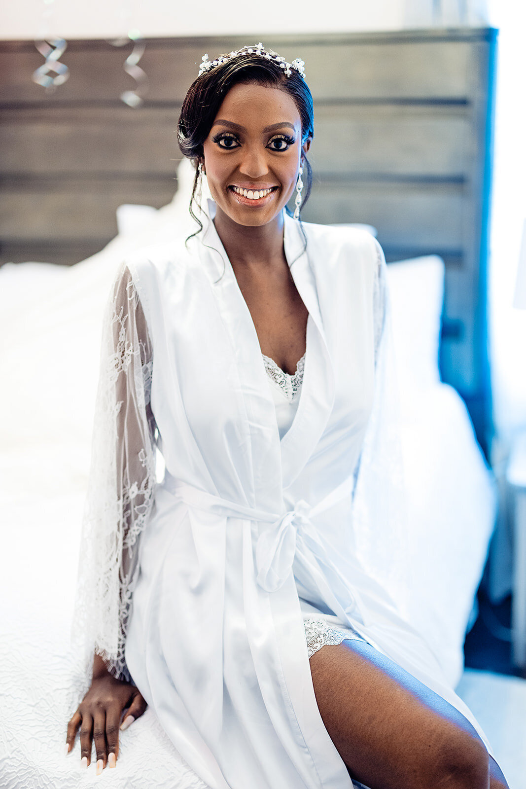 A bride in a white robe with lace trimmings sits on a bed, looking up and smiling, her hair styled up with a crystal headpiece