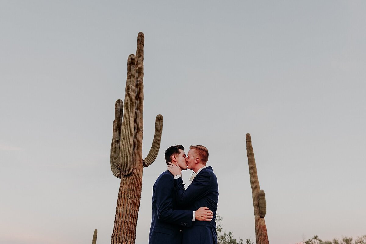 Two grooms embrace and kiss surrounded by saguaros  in the desert