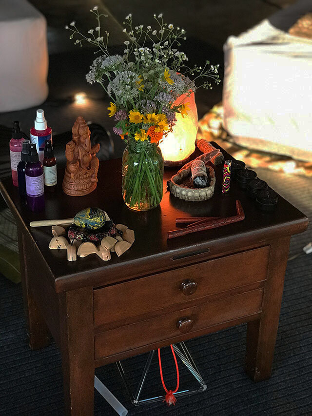 A photo of a ceremonial alter with a ganesha statue, cleansing sprays, flowers, smudging tools, a hapeh pipe, and rattle.