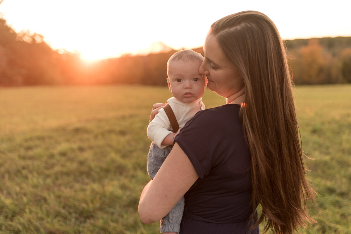 Mother is pressing her nose against infant son's forehead as the sun sets behind them in a green field