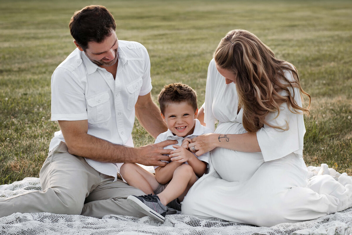 dad and mom tickling their young son wearing white outfits sitting on a white blanket in the grass in the summer time