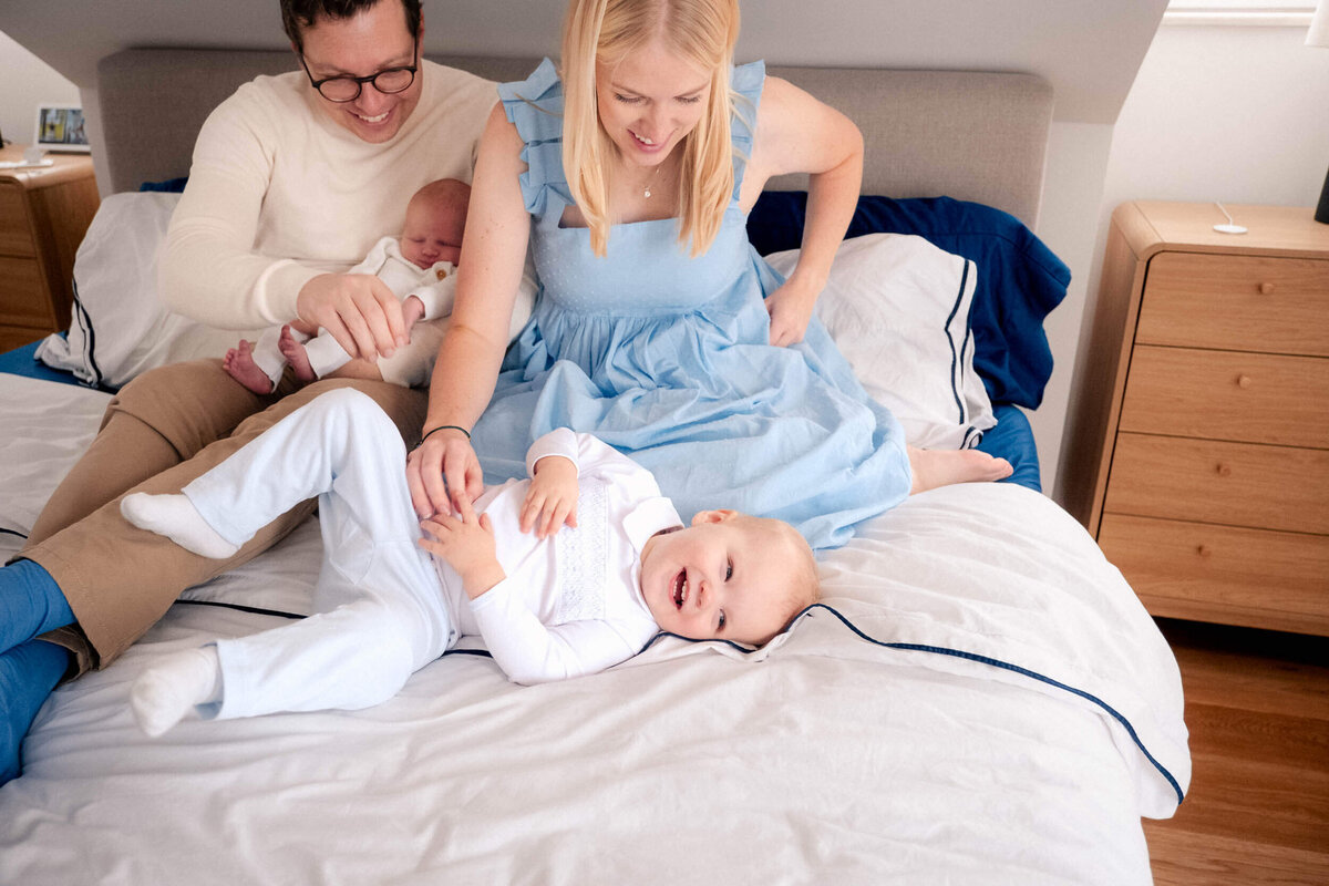 Young family on a bed, with mother and father smiling as toddler giggles and newborn is cradled.
