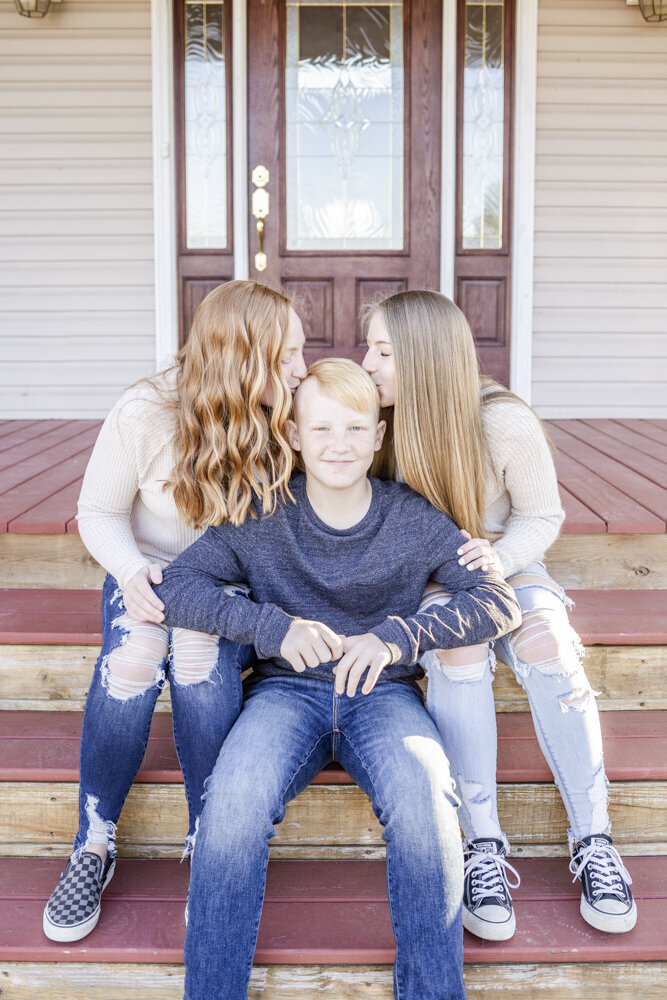 Two sisters kiss brothers head on their front porch while brother smiles in Georgetown, Ohio.