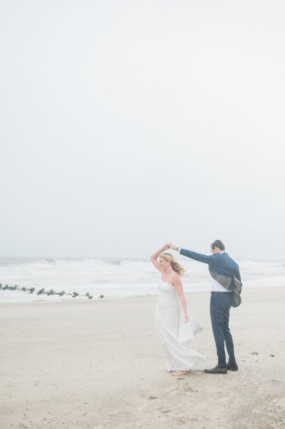 The bride laughs as the groom lifts her arm for a twirl, their feet sinking into the wet sand