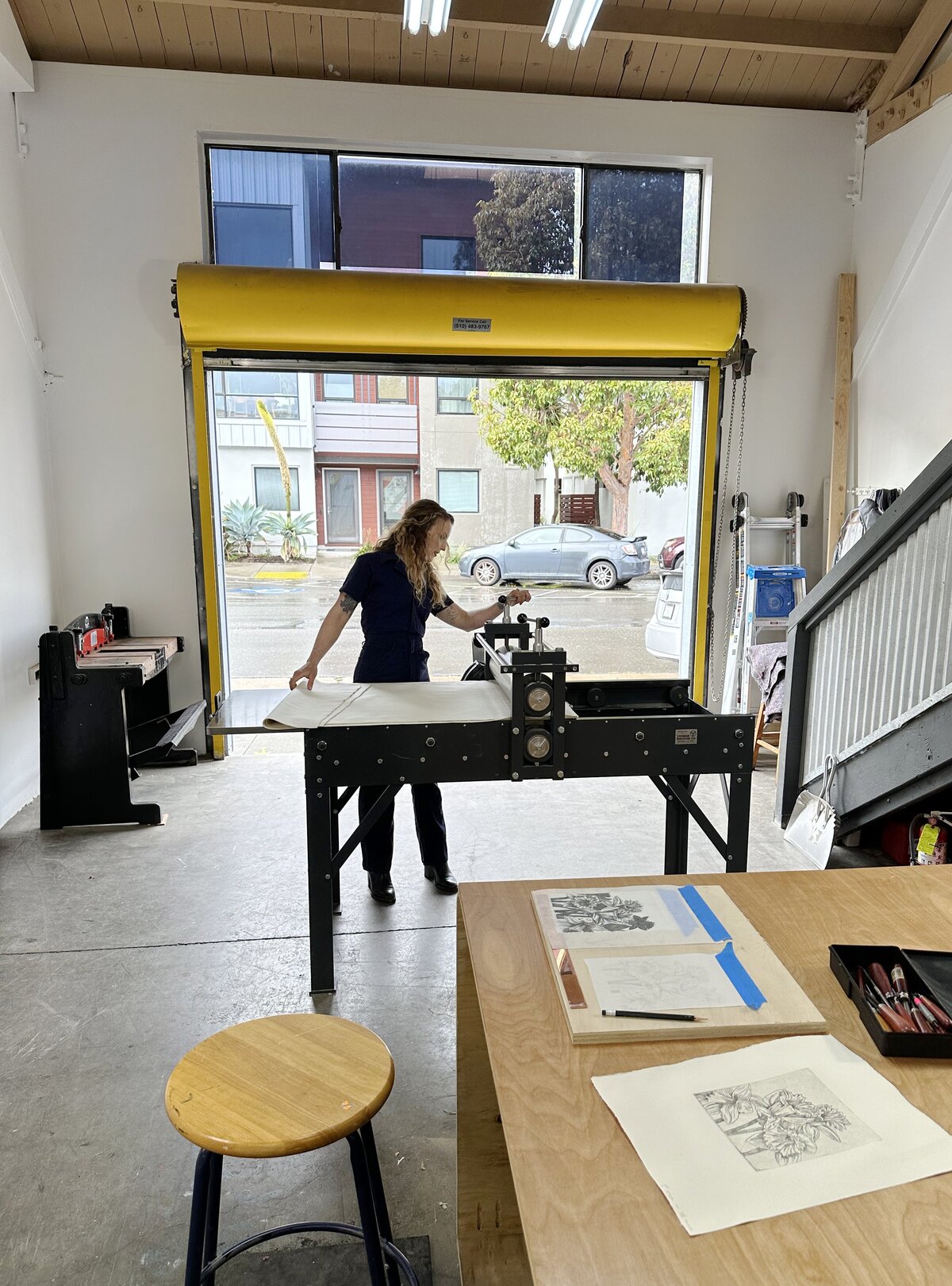 A master printer operating a professional rolling press machine in a brightly-lit printmaking studio with large windows.