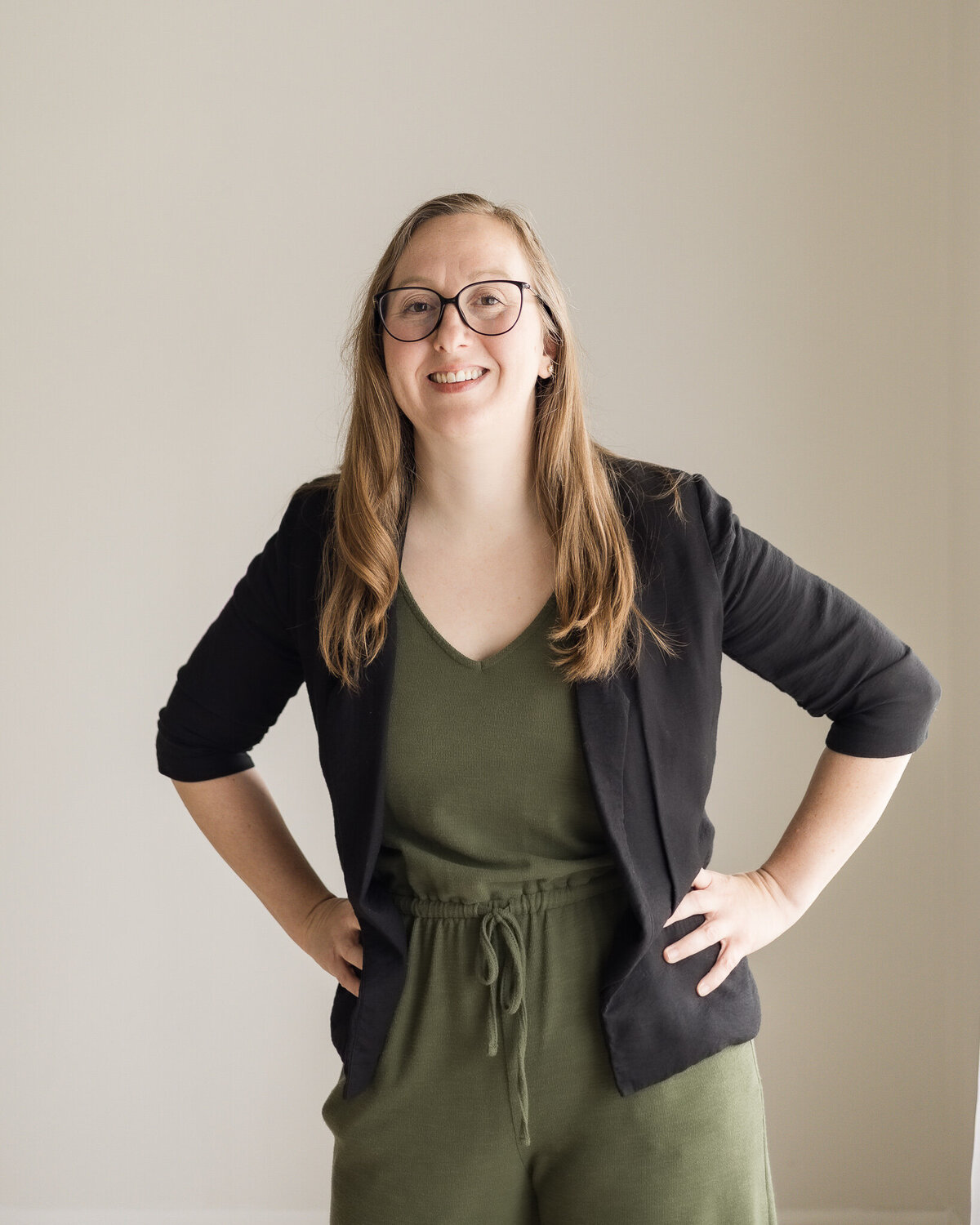 Strawberry blonde female, standing with her hands on her hips, wearing a dark green shirt and pants, with a black jacket over the green outfit, wearing glasses, smiling at the camera