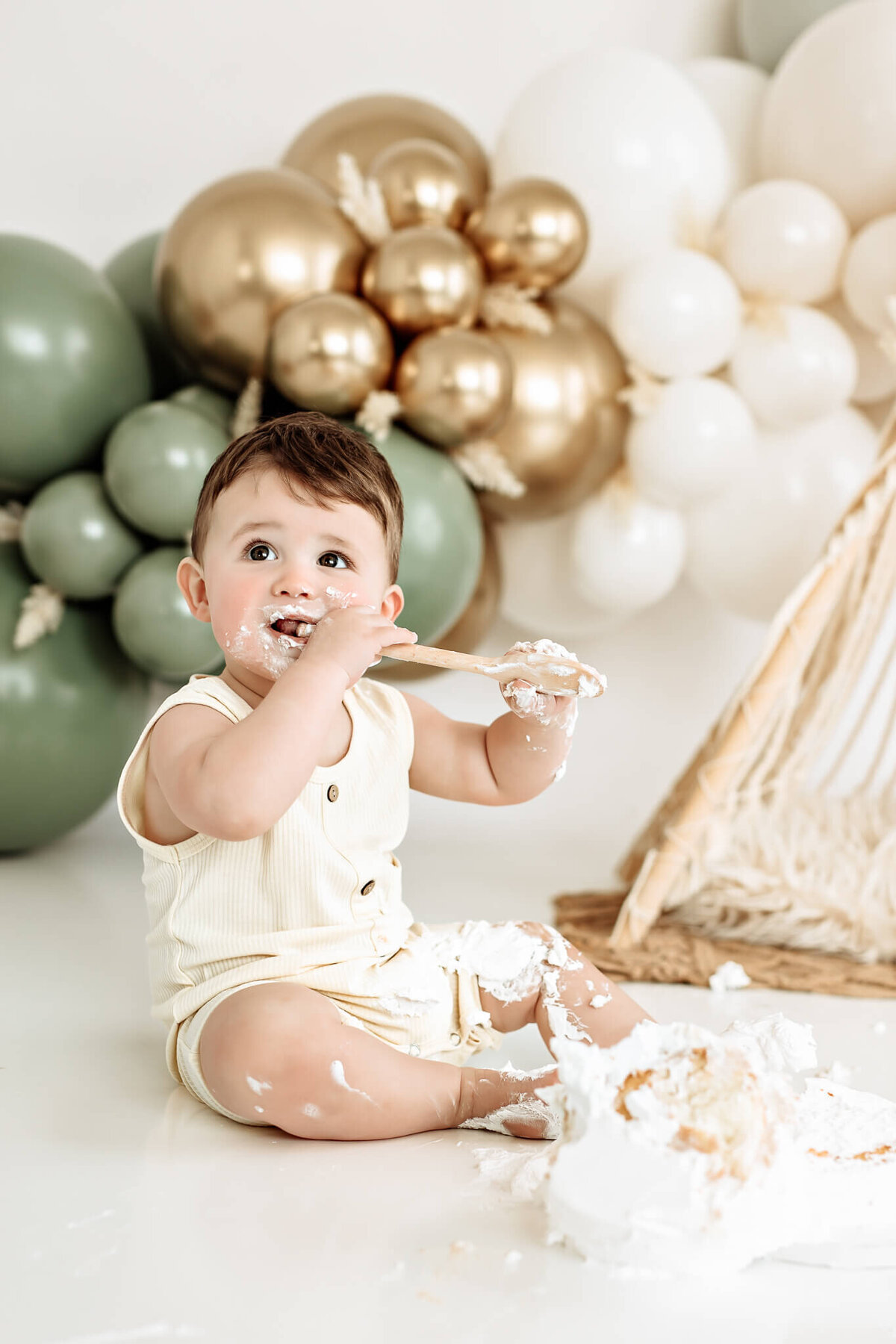 A baby sits on the floor, eating cake with a wooden spoon. The scene is decorated with green, gold, and white balloons in the background. The baby, wearing a light-colored outfit, is playfully covered in cake and frosting.