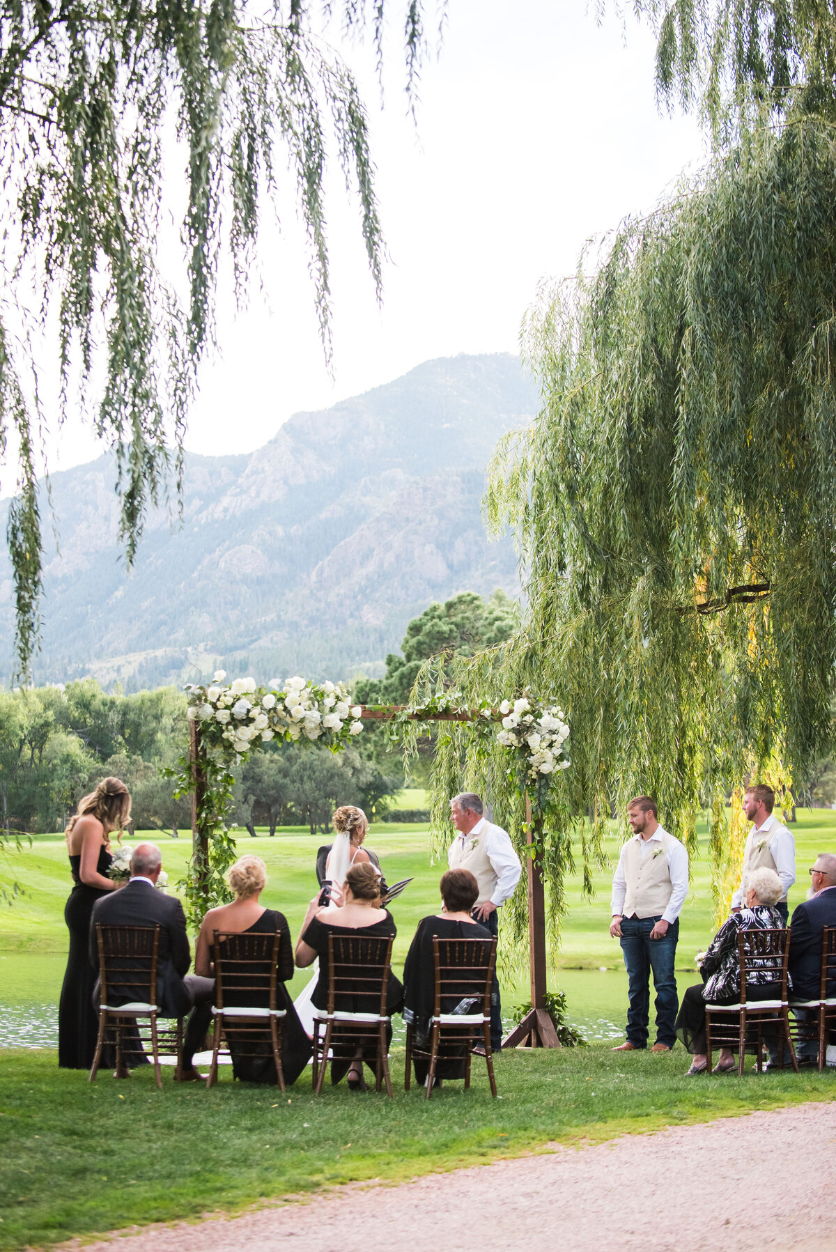 A wide angle shot of a ceremony under a floral wedding arch next to a weeping willow