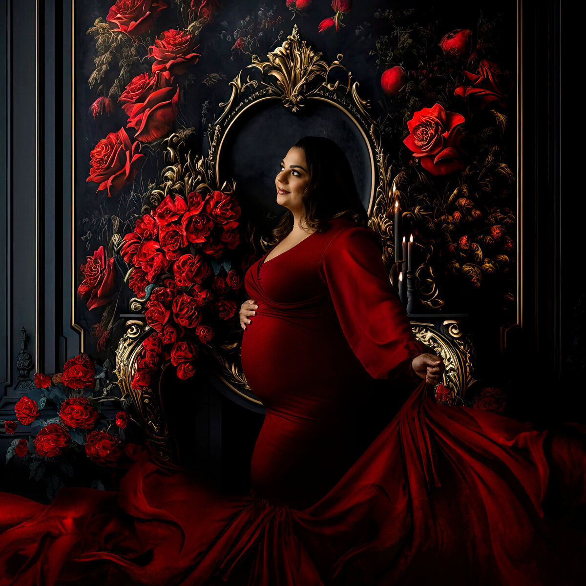 An expecting mother in a long flowing red maternity gown stands in a studio with a wall of roses behind her