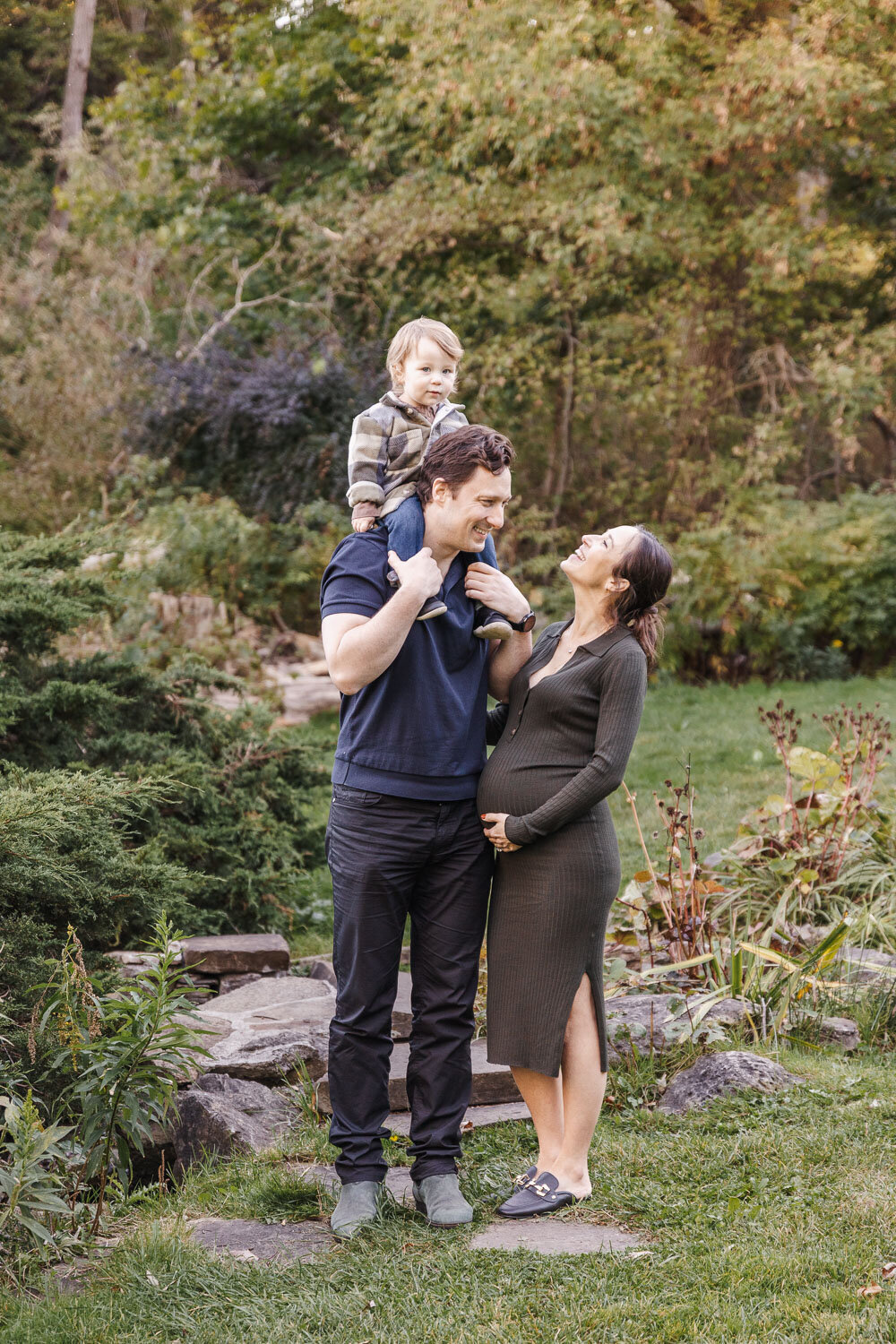 Toddler boy sitting on dads shoulders while pregnant mom lovingly smiles up at him.