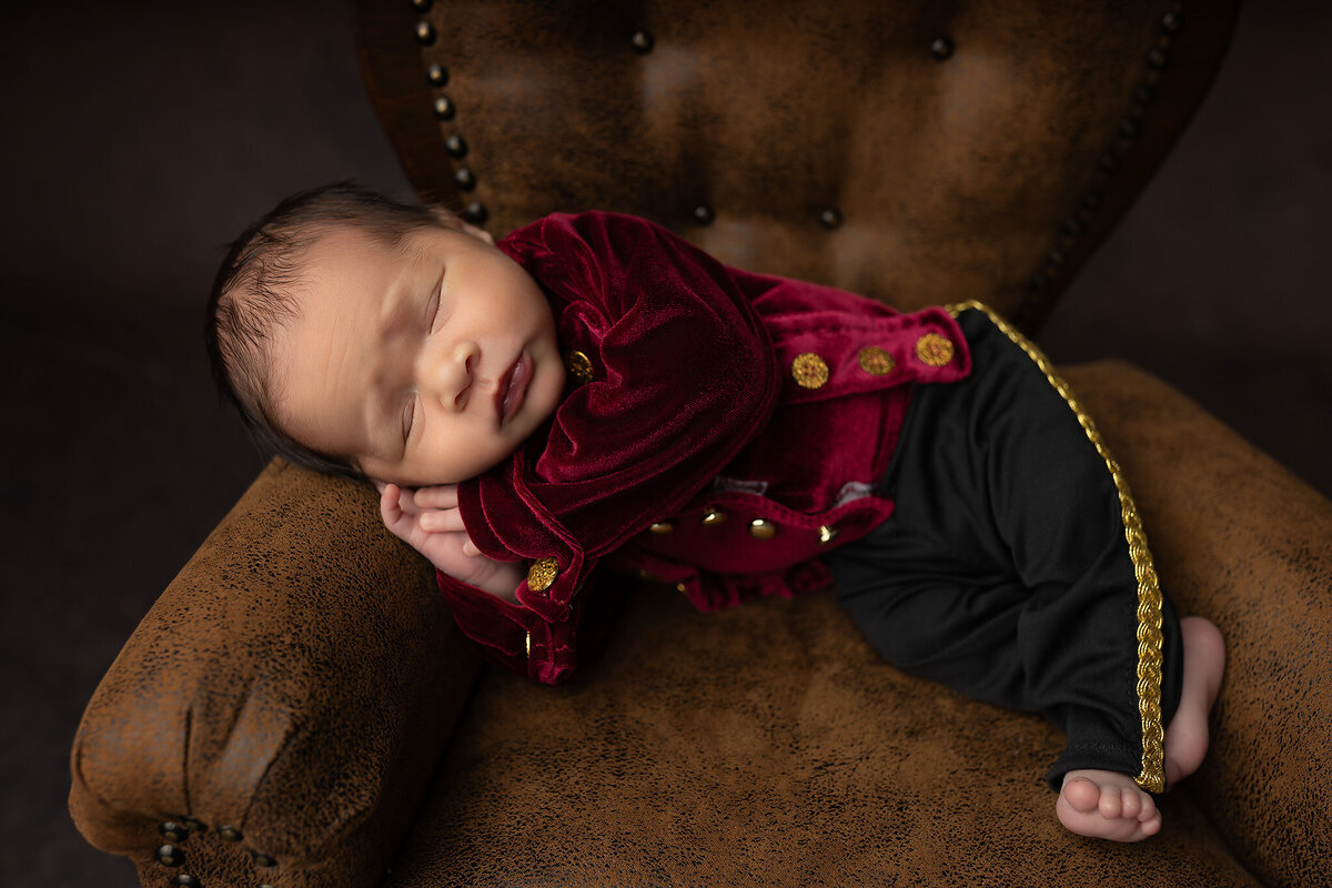 Newborn baby in red velvet jacket and gold accents asleep on chair arm — newborn photography studio portrait.