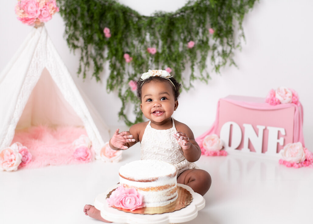 Baby photoshoot for first birthday — smiling one-year-old girl in lace romper with naked cake and floral greenery backdrop