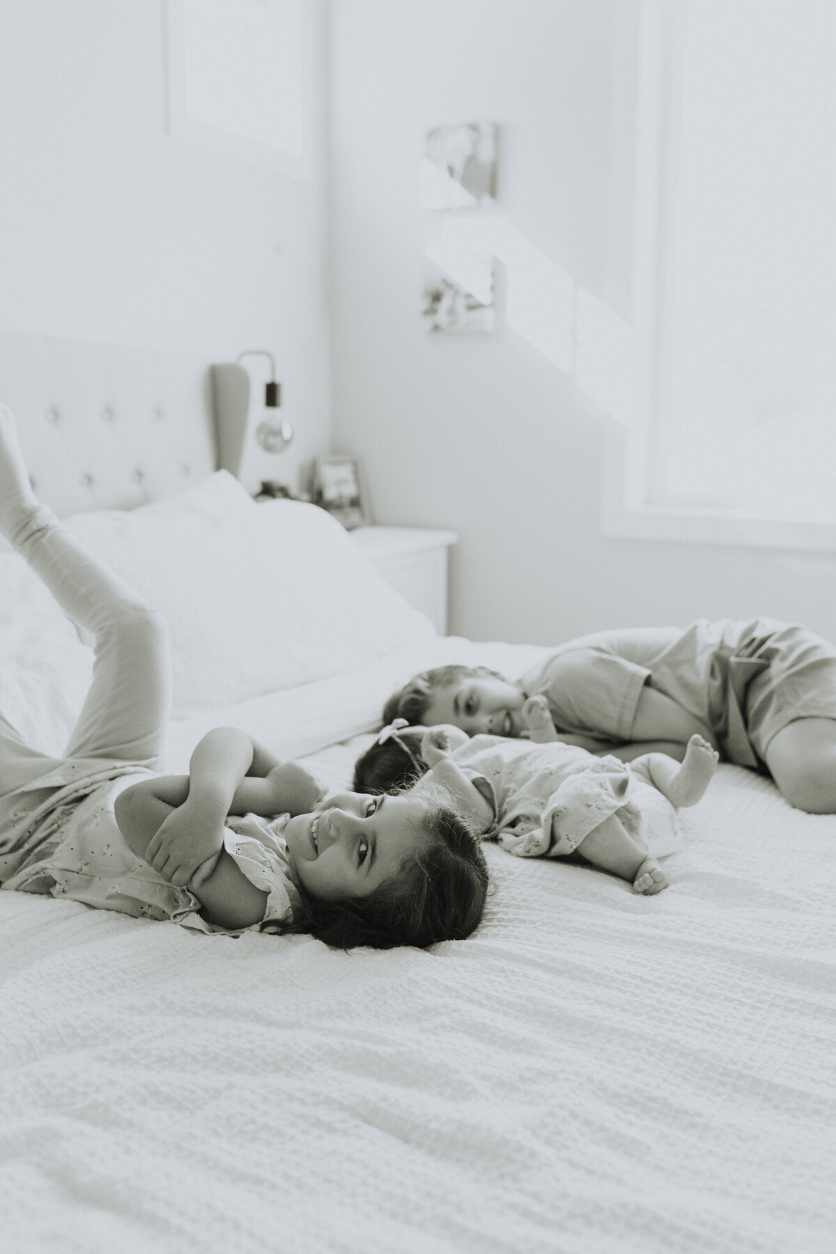 black and white photo of a young boy and girl laying on the bed beside their baby sister while the older girl looks at the camera smiling taken by a langley family photographer