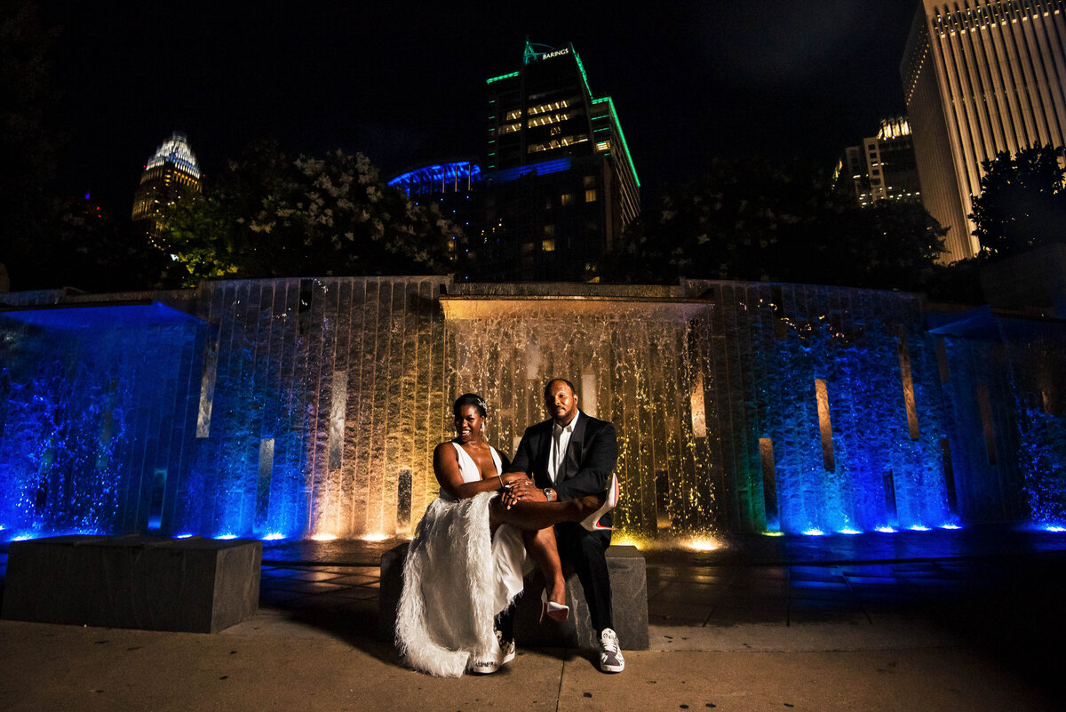 Portrait of black couple who just eloped sitting in Romare Bearden Park  in front of the blue and yellow waterfall by Charlotte wedding photographer DeLong Photography