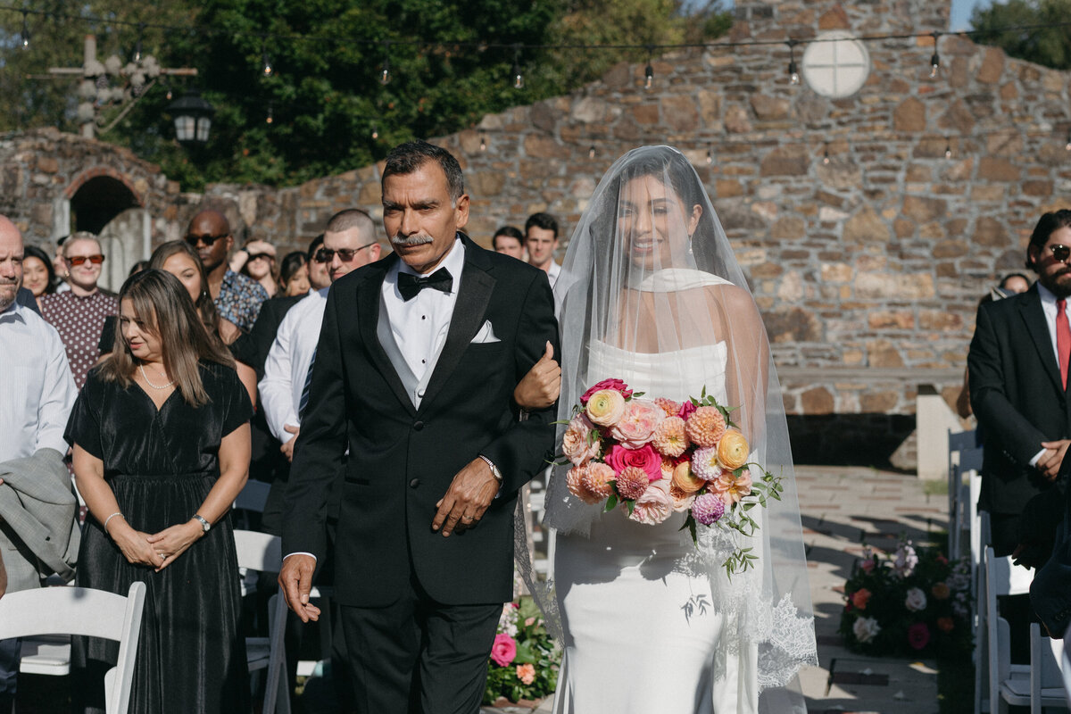 Bride walking down the aisle holding a vibrant bouquet of roses, dahlias, and ranunculus in pink, peach, and cream tones, surrounded by guests at an outdoor stone chapel ceremony.
