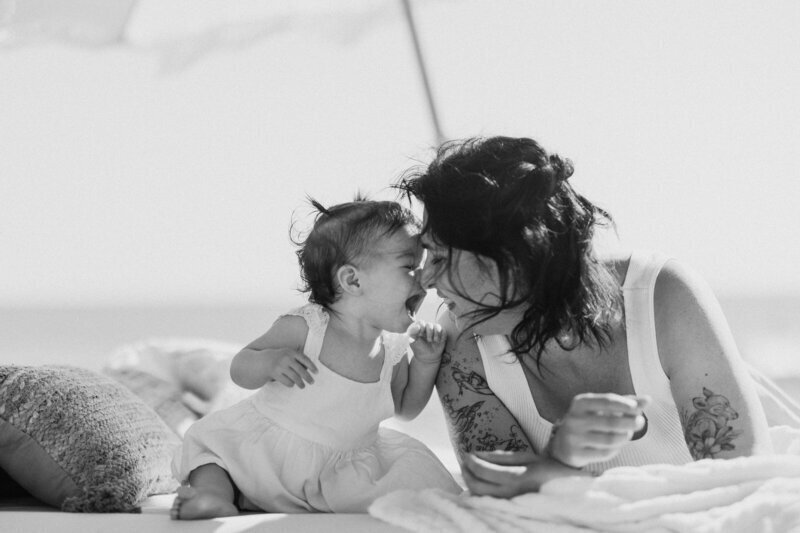 Photo capturée en noir et blanc par Laura, photographe professionnelle  en Vendée, d'une maman allongée sur le ventre complice avec sa petite fille dans un décor cosy installé sur la plage.