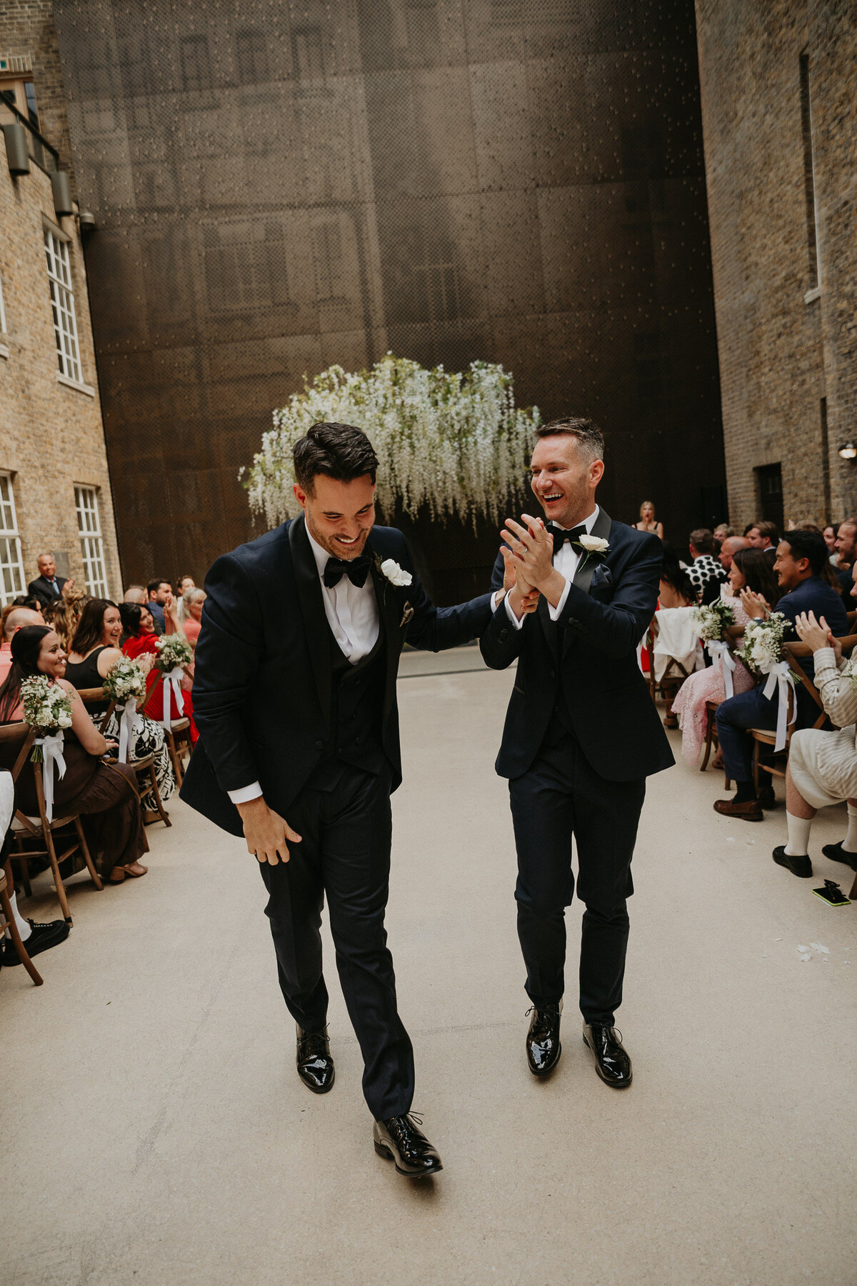 Two grooms walk back up the aisle as their guests cheer at Hackney Town Hall. 