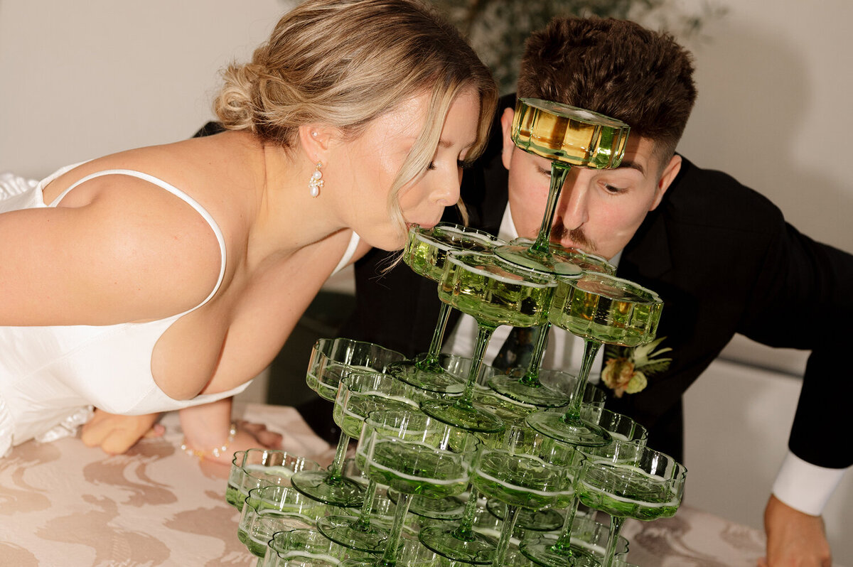 A bride and groom lean over a champagne tower, both sipping from green glassware.