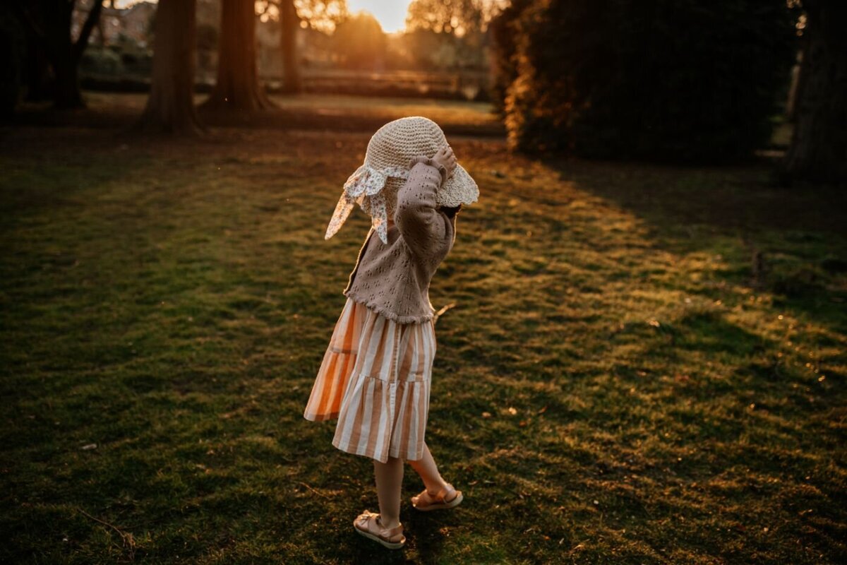 a little girl with curly hair wearing a sun hat, walking across a park at sunset with trees silhouetted in the background.