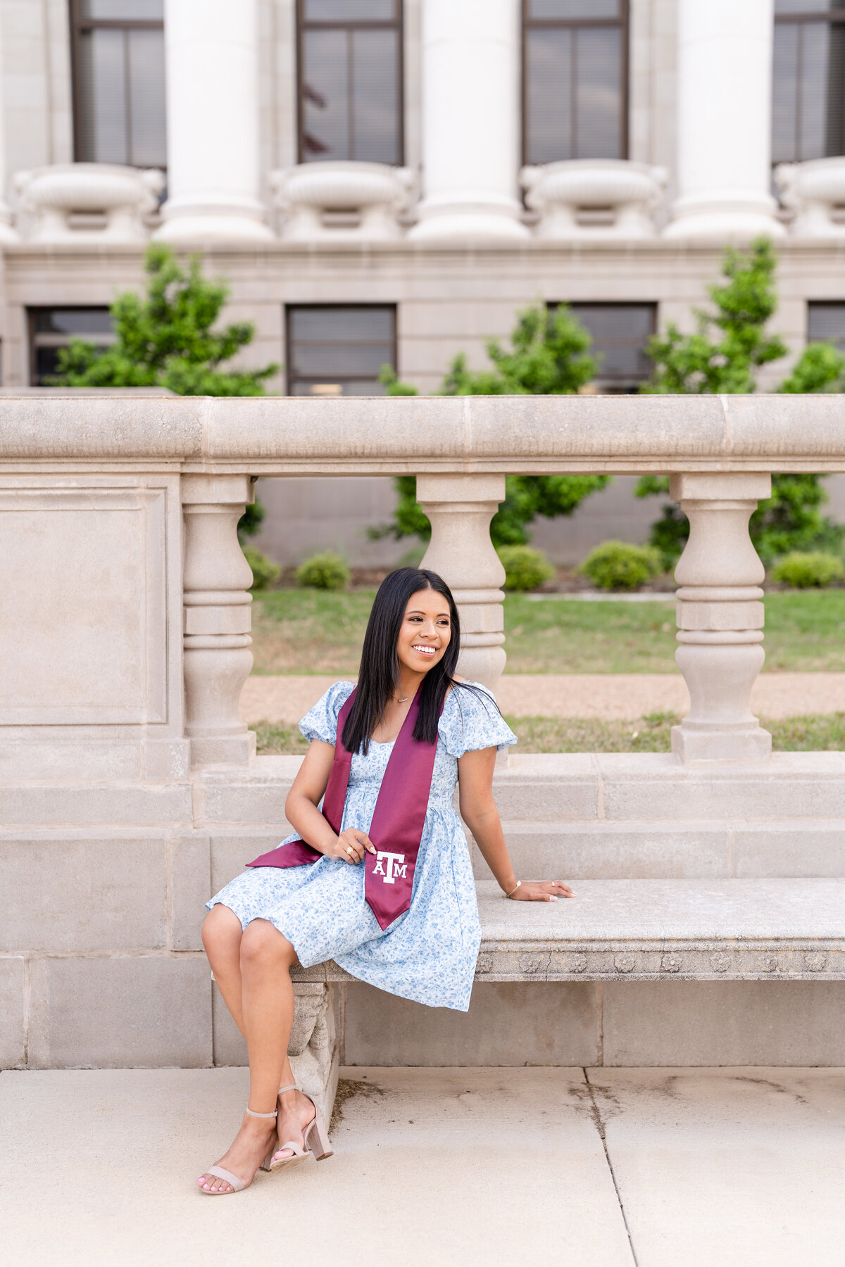 Texas A&M senior girl sitting on bench and wearing blue dress and maroon stole while smiling away in front of Administration Building