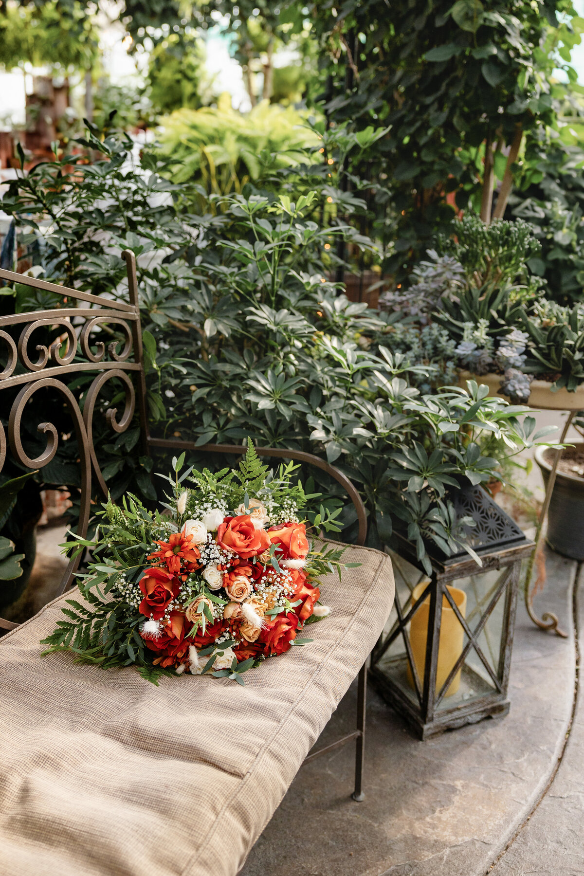 A vibrant bridal bouquet featuring orange and peach roses, white flowers, and lush greenery rests on a beige cushioned bench in a garden setting.