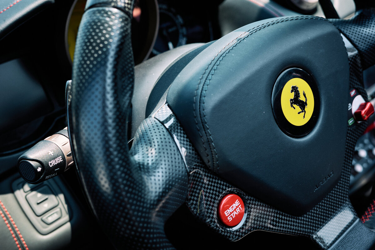 Close-up of a Ferrari’s carbon fiber steering wheel featuring the iconic yellow Ferrari emblem and red engine start button, showcasing the luxurious interior detailing.