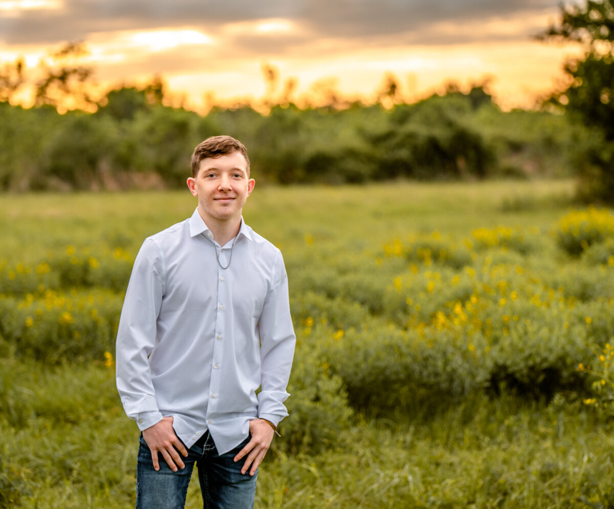 A senior boy has his hands in his pockets while standing in a field of bushes and yellow flowers.