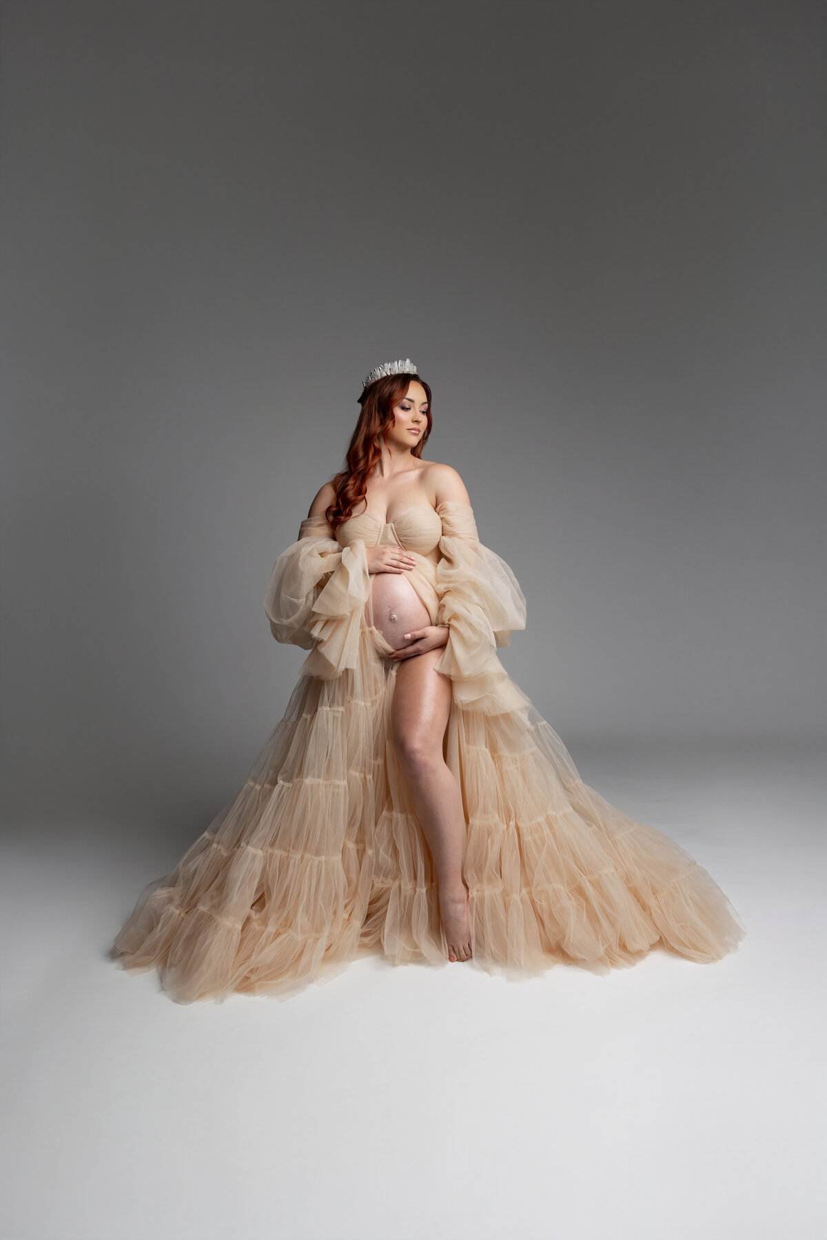 The expectant mother, wearing a yellow dress and a crown, gently tilts her head while cradling her belly. Captured by Inland Empire maternity photographer Kaitlyn Dawn Photography.