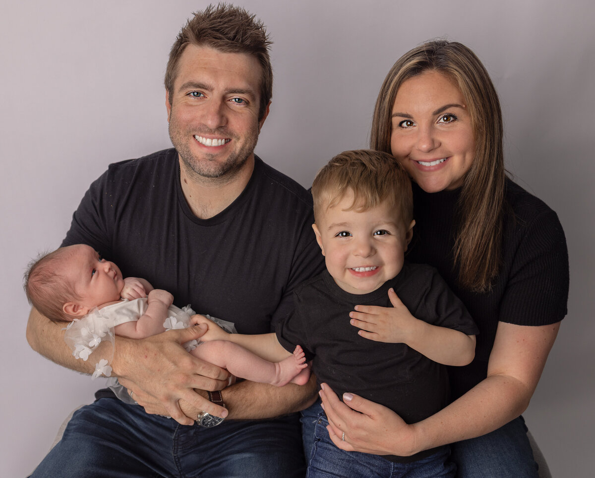 Smiling family of four during a newborn photo session in Waldwick NJ with parents holding their baby girl and toddler son