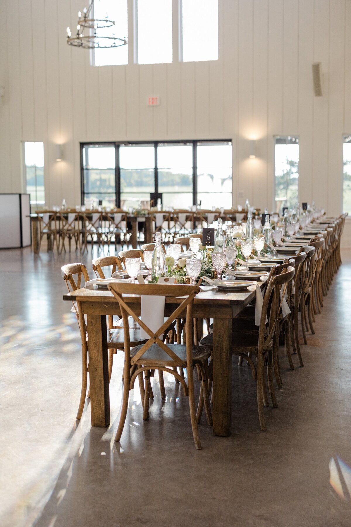 Detail shot of a reception table at a wedding reception at Bella Cavalli Evenst in Aubrey, Texas. The long, wooden, rectangular table is covered by many place settings, glasses, candles, and floral arrangements.  similar table can be seen in the background.