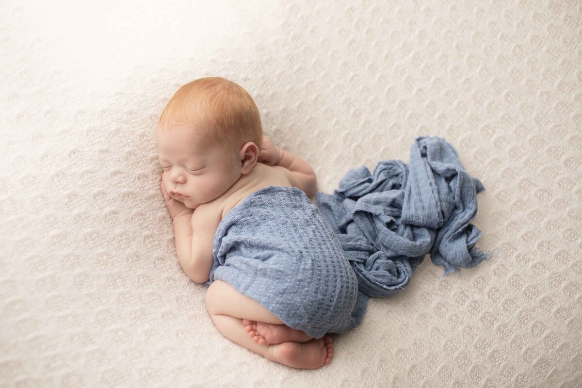 Tiny newborn boy with soft red hair posed with his hands under his chin on a dreamy fabric backdrop